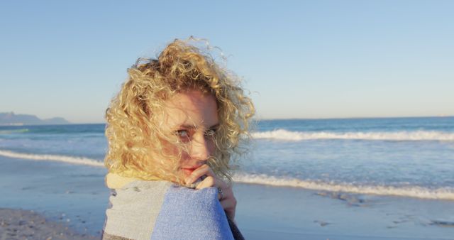 Blonde woman with curly hair wrapped in towel on a beach. She looks over her shoulder towards the camera as waves gently wash ashore. Ideal for travel ads, summer vacation promotions, lifestyle blogs, or nature magazines.