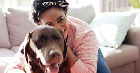 African American woman cuddling with a brown Labrador exemplifies the affectionate bond between pets and their owners. Natural sunlight enhances the warmth and coziness, making it perfect for themes of relaxation, companionship, and home life. Suitable for advertising pet products, depicting family-friendly home environments, or illustrating articles about pet-owner relationships.