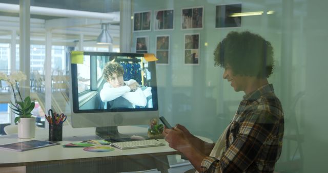 Man with curly hair and plaid shirt sitting at desk in modern office, texting on smartphone while computer is on desk. Concept of work-life balance, productivity, office culture, and modern work environments. Ideal for business, technology, and office-related content.