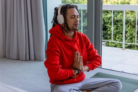 African american man meditating with headphones in bright living room near balcony