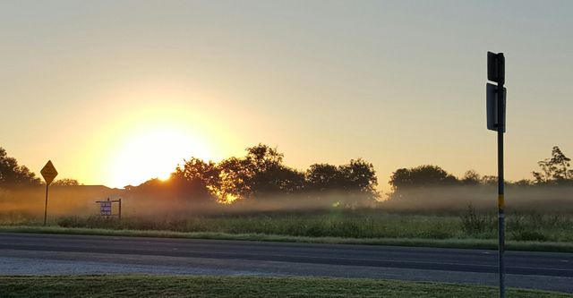 Sunrise over a mist-covered field next to a quiet road. Vibrant morning colors with the sun creating a hazy, calm atmosphere. Ideal for depicting serenity, rural living, or scenic beauty in nature. Perfect for backgrounds, blogs on peace and tranquility, or travel and nature websites.