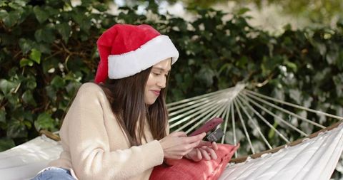 Young woman wearing santa hat relaxing on hammock while shopping online with credit card