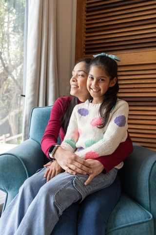 Indian mother embracing daughter on teal armchair by window, sunlit cozy family bonding