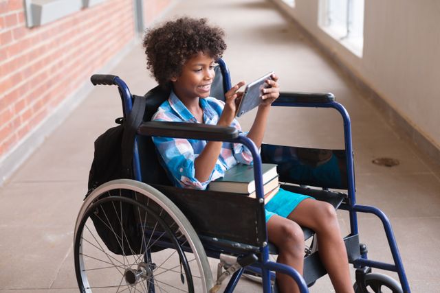 Smiling african american elementary boy using digital tablet while sitting on wheelchair in corridor. unaltered, education, disability, wireless technology, physical disability and school concept.