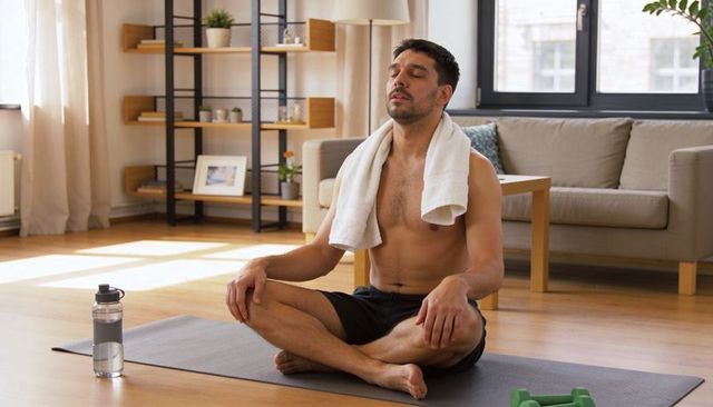 Shirtless man meditating on yoga mat in bright living room cooling down after workout