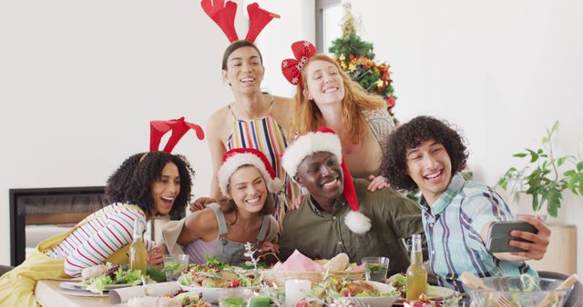 Group of friends from diverse backgrounds enjoying a joyful Christmas celebration around a festive meal. Smiling and laughing while posing for a selfie, with some wearing Santa and reindeer hats. Ideal for promoting holiday gatherings, festive joy, and the spirit of togetherness. Perfect for advertisements, blog posts, and social media campaigns centered on Christmas celebrations, friendship, and multicultural events.