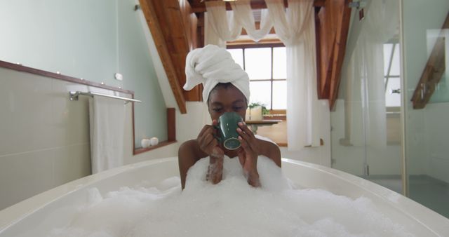 African american attractive woman relaxing in foam bath and drinking coffee in bathroom. beauty, pampering, home spa and wellbeing concept.