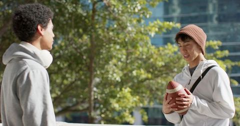 College friends chatting on urban campus holding football wearing hoodies and beanie