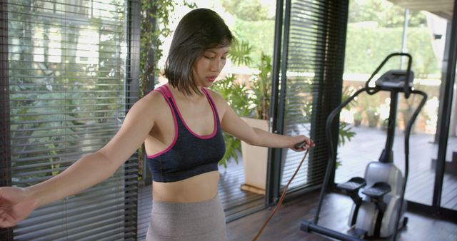 Focused asian woman holding skipping rope in sunny home gym. Healthy lifestyle, fitness and wellbeing, exercises, unaltered.