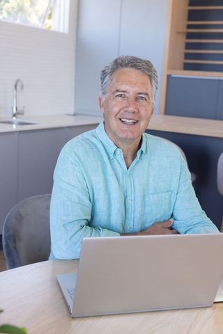 Mature man working remotely at laptop in modern minimalist kitchen smiling and productive