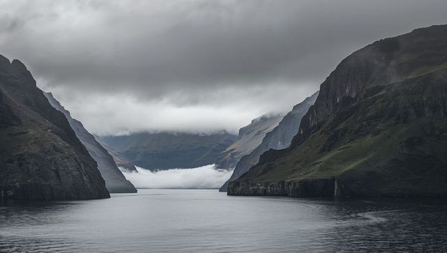 Misty fjord inlet with calm reflective water, dramatic cliffs and low-hanging clouds