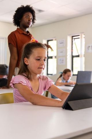 Elementary student using tablet while teacher supervising classroom learning and focus