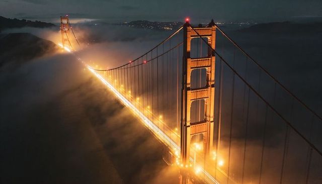Golden gate bridge glowing and piercing thick fog at night with amber light trails