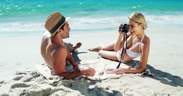 Couple enjoying a sunny day on the beach. Woman in swimsuit takes photos with camera while man in hat smiles. Perfect for travel blogs, vacation advertisements, summer getaway promotions, and photography-related content.