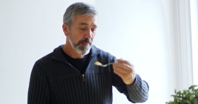 Senior man with gray hair and beard eating a healthy meal with a fork. He is wearing a casual dark outfit and appears thoughtful while enjoying his food. Ideal for use in articles or advertisements related to senior nutrition, healthy eating habits, or lifestyle topics.
