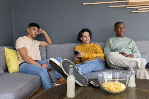 Three diverse young men relaxing on gray sectional sofa with chips and bottled drinks
