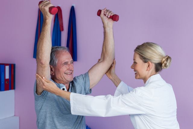 Senior man receiving assistance from a physiotherapist while lifting dumbbells in a clinic. Ideal for use in healthcare, physical therapy, rehabilitation, elderly fitness, and wellness-related content.