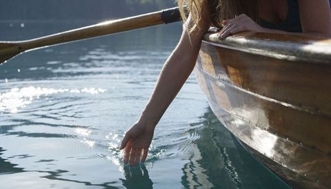 Leaning woman trailing hand in calm lake from varnished rowboat, sunlight reflections
