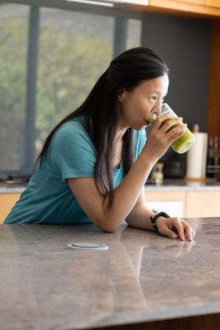 Mature asian woman leaning on marble counter drinking green smoothie in modern kitchen