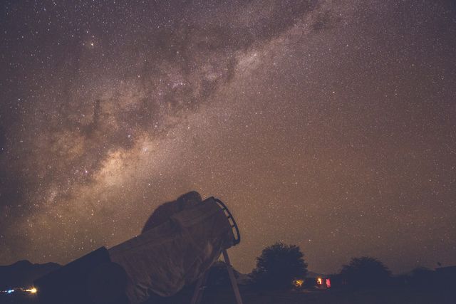 Person using a large telescope for stargazing under densely-filled night sky. Milky Way and various stars are clearly visible. Useful for illustrating topics related to astronomy, space exploration, night photography, or science education.
