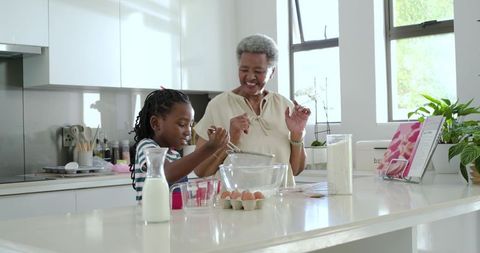 Grandmother teaching granddaughter baking and whisking batter in bright modern kitchen