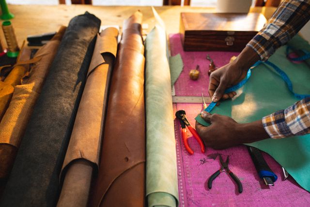 Cropped hands of african american young craftsman measuring leather at workbench in workshop. unaltered, small business, craftsmanship, handcraft, leather craft and workshop.