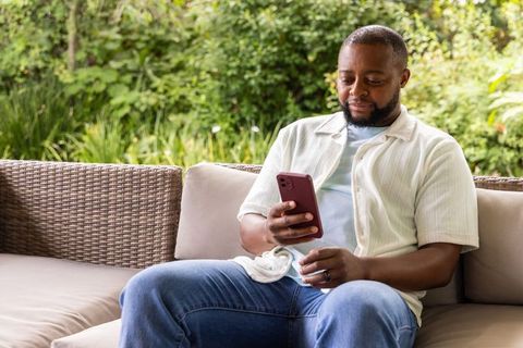 Relaxed african american man using smartphone on outdoor rattan sofa with face mask