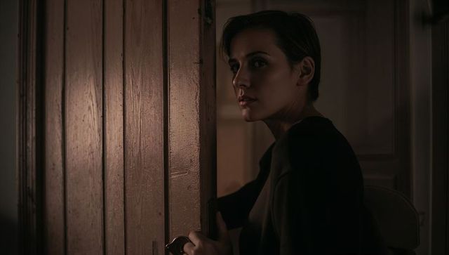 Woman peering through partially open paneled door holding doorknob, moody interior lighting