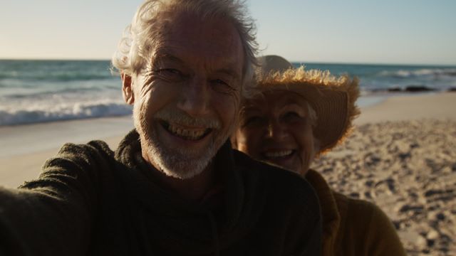 Senior couple enjoying a relaxed moment on the beach, smiling and taking selfies with the ocean in the background. Perfect for themes of retirement, happiness in later life, beach holidays, and outdoor lifestyle. Suitable for advertisements, greeting cards, travel brochures, and articles on senior well-being.