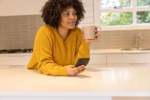 Relaxed woman sipping coffee and scrolling smartphone on kitchen island, modern lifestyle