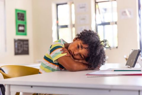 African american schoolboy resting head on arms at classroom desk with tablet, looking contemplative