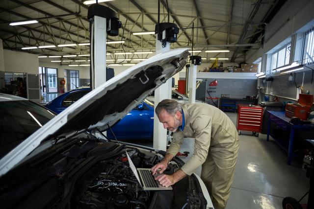 Mechanic using laptop while servicing a car engine in repair shop