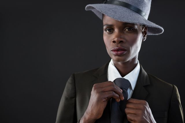 This image depicts an androgynous man adjusting his tie while wearing a stylish suit and hat against a grey background. Ideal for use in fashion editorials, advertisements, and articles focusing on modern style, gender fluidity, and professional attire. Perfect for illustrating concepts of confidence, sophistication, and contemporary fashion trends.