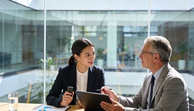 Senior executive mentoring junior colleague reviewing tablet and charts in glass office