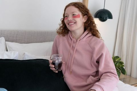 Smiling redhead woman relaxing on bed in pink hoodie holding glass mug with dark beverage