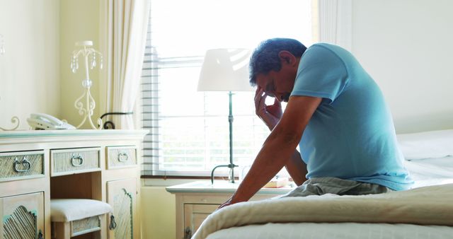 A middle-aged man appears distressed or deep in thought while sitting on the edge of a bed in a well-lit bedroom, with copy space. His posture and expression suggest a moment of sadness, worry, or exhaustion.