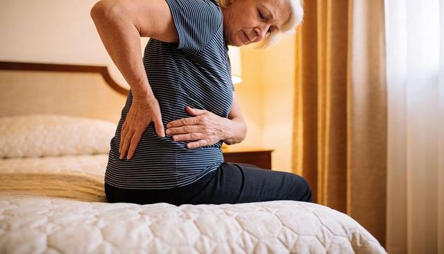 Senior woman pressing lower back while sitting on bed experiencing back pain