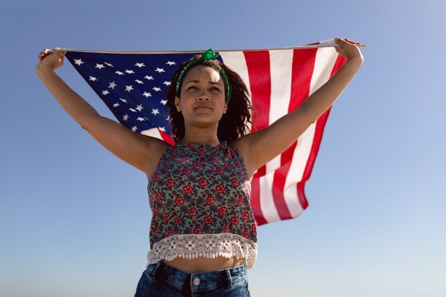 Biracial woman holding American flag, standing against sky. She has brown skin, curly dark hair, and is wearing casual clothes, unaltered