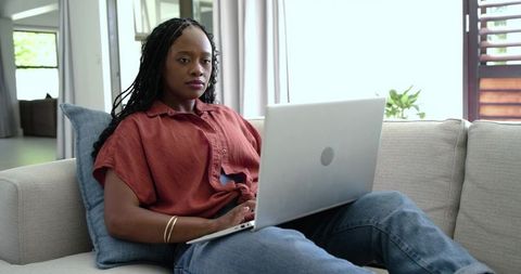 Remote woman working on laptop on sofa in modern sunlit minimalist living room