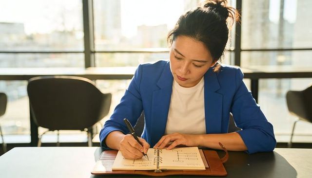 Professional woman wearing blue blazer writing leather-bound planner at sunlit coworking table