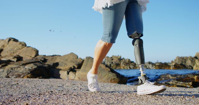 Individual with a prosthetic leg walking on a rocky beach during a sunny day. Suitable for topics like resilience, outdoor activities, inclusivity, and positive attitudes towards disability. Can be used in websites, promotional materials, blogs, or social media posts about independent living, adaptive sports, or inspirational stories.