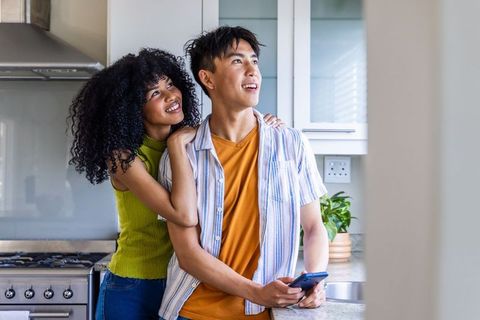 Young multicultural couple embracing in bright kitchen, holding smartphone and smiling