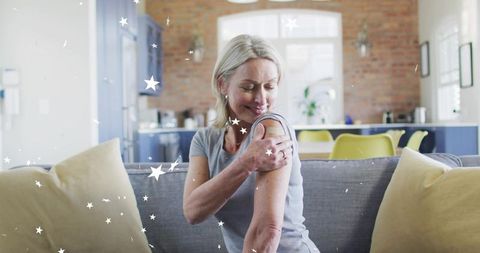 Mature woman touching upper arm with bandage while relaxing on sofa in sunlit kitchen