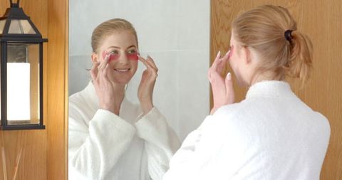 Woman wearing bathrobe applying gel eye masks in modern bathroom