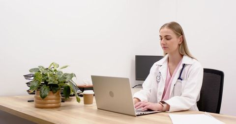 Female clinician typing on laptop at minimalist clinic desk with stethoscope and monitor