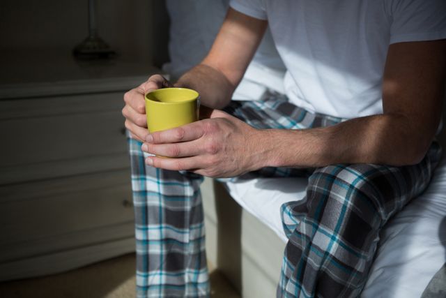 Man sitting on bed in bedroom, holding yellow coffee mug, wearing plaid pajamas. Ideal for themes of morning routines, relaxation, home comfort, casual lifestyle, and personal time. Suitable for use in blogs, articles, advertisements, and social media posts related to daily routines, home life, and relaxation.