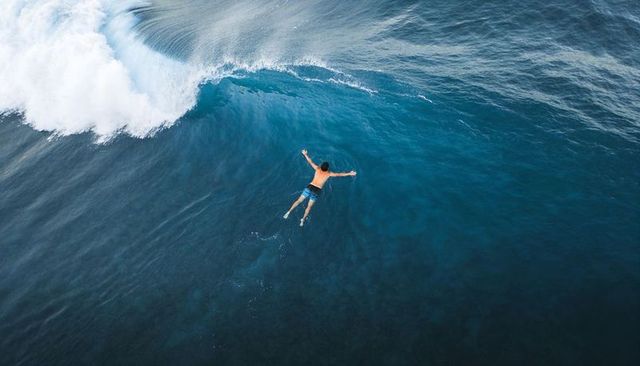 Aerial view of lone swimmer bracing against breaking wave in blue ocean under sunlight