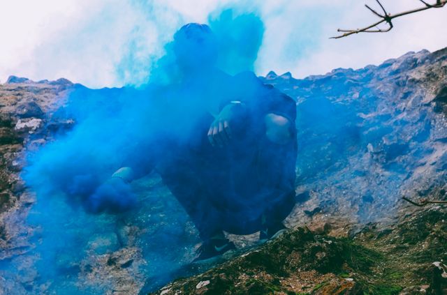 Person covered in dense blue smoke against a rugged rocky background, creating a mysterious and dramatic atmosphere. Useful for themes about mystery, adventure, outdoor exploration, and environmental elements.