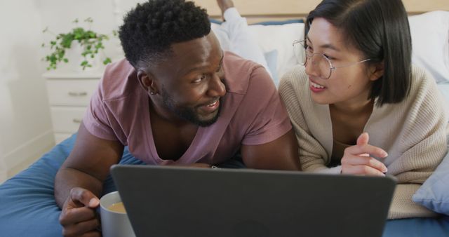 Young multiracial couple lying on bed using laptop, enjoying time together. They seem engaged and happy, signifying a strong bond. Could be used for themes like modern relationships, technology, home life, or diversity and inclusion in everyday scenarios.