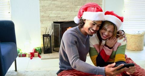 Couple Laughing while Sharing Smartphone on Living Room Floor Wearing Santa Hats